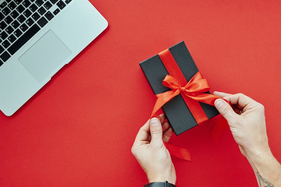 Close-up of hands tying a red ribbon on a gift box beside a laptop on a red background, perfect for holiday presents.